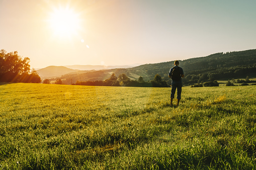 Young man with backpack walking on meadow in summer Czech landscape at sunset
