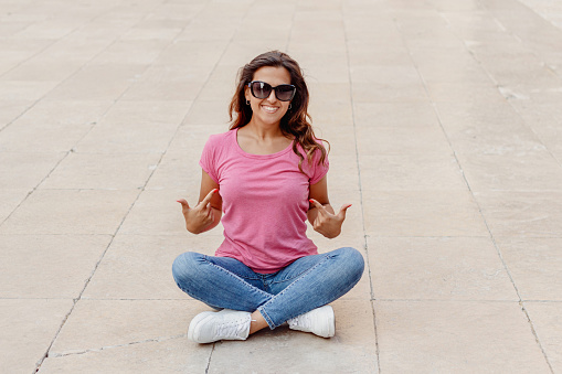 Young women wearing t-shirt and jeans sits on the pavement and shows in the  center