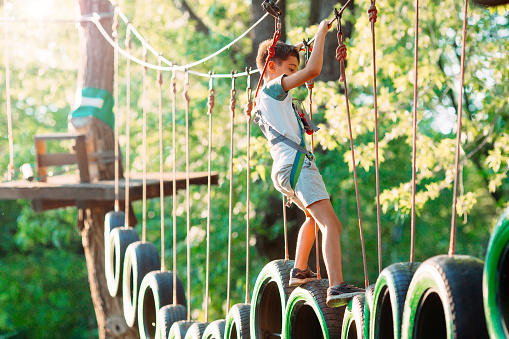 Strong excited young boy playing outdoors in rope park. Caucasian child dressed in casual clothes and sneakers at warm sunny day. Active leisure time with children concept.