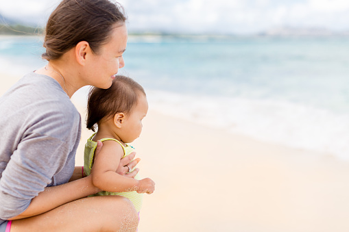 Young female parent and her child daughter watching the ocean coast water waves while sitting on the sand during summer vacations.
