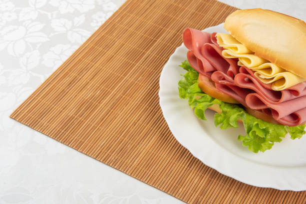Mortadella sandwich, lettuce and cheese on a white plate and a bamboo mat with white background on a table with white towel, Selective focus.