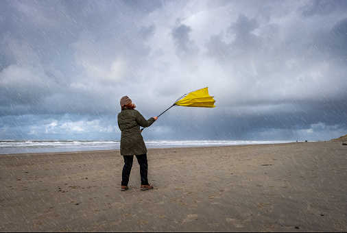 Woman struggling with big yellow umbrella on beach in stormy weather