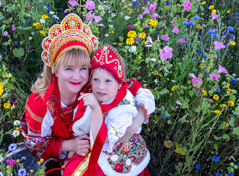Mom 40 years old and daughter 5 years old in national Russian costumes, red sundress and kokosheik. Over the edge of the flower meadow and forest. Summer day.