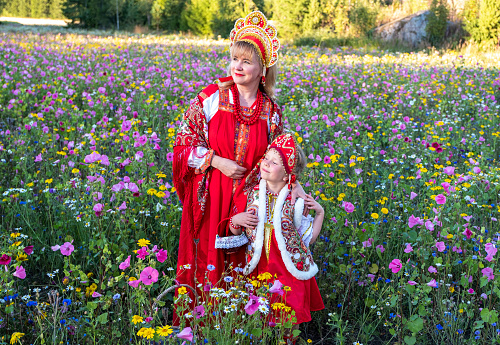 Mom 40 years old and daughter 5 years old in national Russian costumes, red sundress and kokosheik. Over the edge of the flower meadow and forest. Summer day.