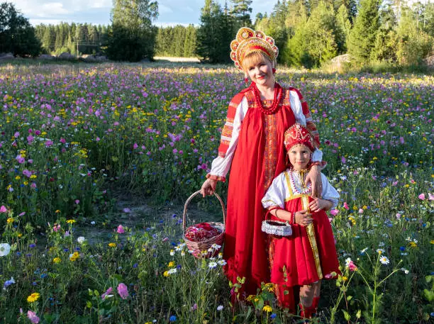 Photo of A family of Caucasians in national Russian costumes. Woman and girl in red sundresses and kokoshniks. Against the backdrop of a rural landscape.