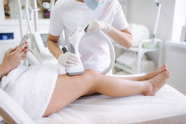 istock Beutiful bussines woman using phone while cosmetologist in white gloves performing radiofrequency lifting procedure on the stomach. Close up 1269285920