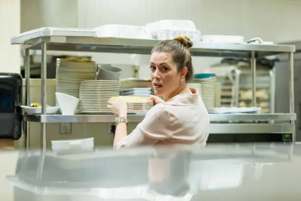A mid adult Hispanic woman in her 30s working in a restaurant. She is in the kitchen, pulling a stack of plates from a shelf.