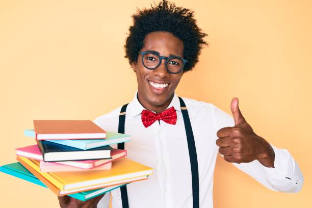 Handsome african american nerd man with afro hair holding books smiling happy and positive, thumb up doing excellent and approval sign Handsome african american nerd man with afro hair holding books smiling happy and positive, thumb up doing excellent and approval sign black nerd stock pictures, royalty-free photos & images