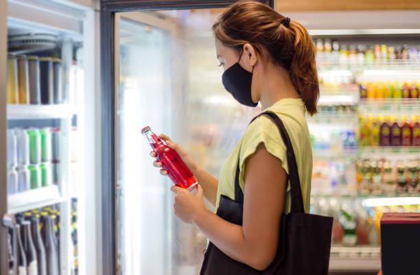 Asian woman reading label in soda bottle before buying Side shot of a young Southeast Asian woman with face mask reading label of the red soda drinks, standing in front of the supermarket freezer  amidst Covid 19 post pandemic convenience store photos stock pictures, royalty-free photos & images
