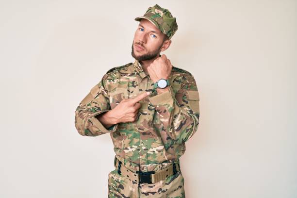 young caucasian man wearing camouflage army uniform in hurry pointing to watch time, impatience, looking at the camera with relaxed expression - clock face clock deadline human hand imagens e fotografias de stock