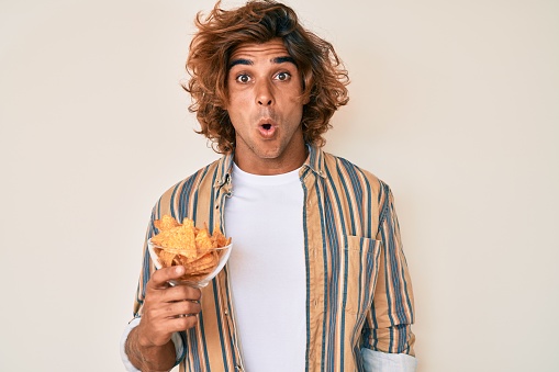 Young hispanic man holding nachos potato chips scared and amazed with open mouth for surprise, disbelief face