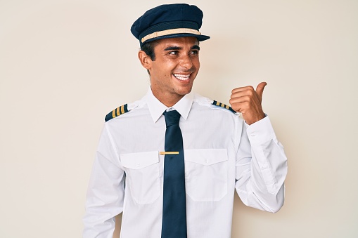 Young hispanic man wearing airplane pilot uniform smiling with happy face looking and pointing to the side with thumb up.