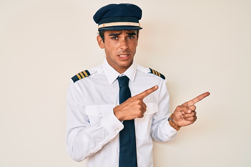 Young hispanic man wearing airplane pilot uniform pointing aside worried and nervous with both hands, concerned and surprised expression