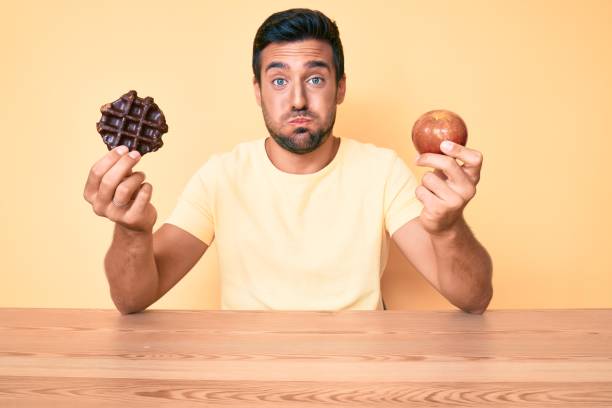 Young hispanic man eating breakfast holding chocolate waflle and apple puffing cheeks with funny face. mouth inflated with air, catching air.