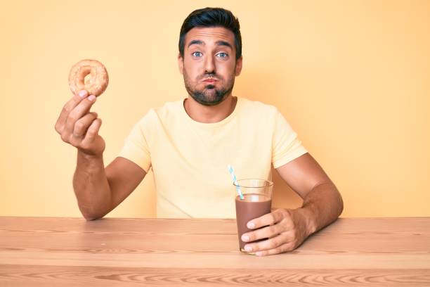 istock Young hispanic man sitting at the table drinking chocolate beverage holding donut puffing cheeks with funny face. mouth inflated with air, catching air. 1269308280
