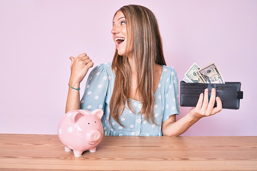 Young blonde girl holding wallet with dollars and piggy bank sitting on the table pointing thumb up to the side smiling happy with open mouth