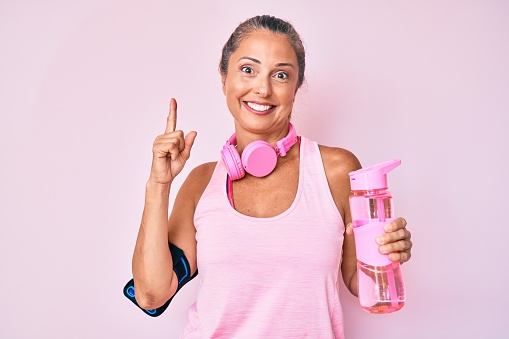 Middle age hispanic woman wearing sportswear holding water bottle smiling with an idea or question pointing finger with happy face, number one