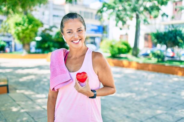 sportiva di mezza età che chiede assistenza sanitaria tenendo il cuore al parco - heart health foto e immagini stock