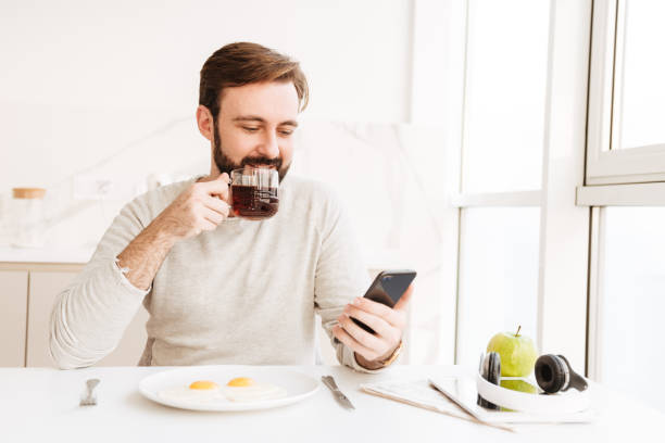 istock Bearded man in casual shirt drinking tea and using smart phone, while having healthy meal with frying eggs at home 1269311385