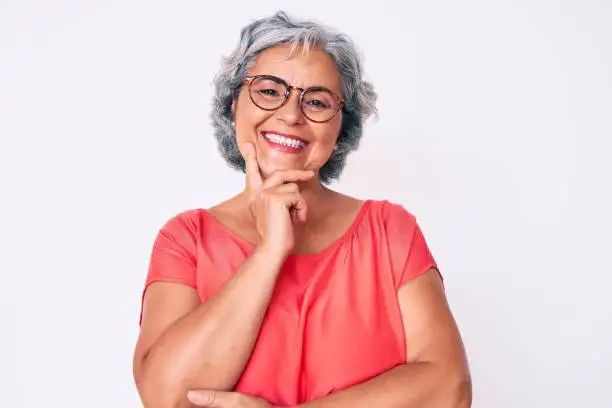 Photo of Senior hispanic grey- haired woman wearing casual clothes and glasses looking confident at the camera with smile with crossed arms and hand raised on chin. thinking positive.