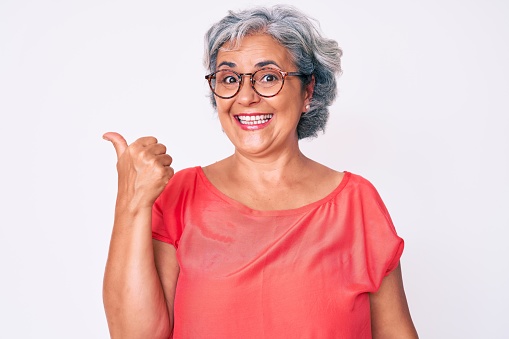 Senior hispanic grey- haired woman wearing casual clothes and glasses smiling with happy face looking and pointing to the side with thumb up.