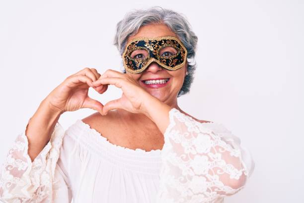 Senior hispanic grey- haired woman wearing venetian carnival mask smiling in love doing heart symbol shape with hands. romantic concept. Senior hispanic grey- haired woman wearing venetian carnival mask smiling in love doing heart symbol shape with hands. romantic concept. carnival mask women party stock pictures, royalty-free photos & images