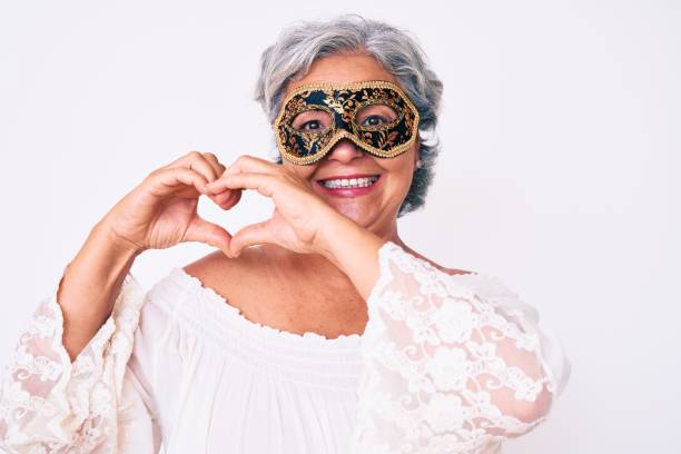 senior hispanic grey- haired woman wearing venetian carnival mask smiling in love doing heart symbol shape with hands. romantic concept. - carnival mask women party imagens e fotografias de stock