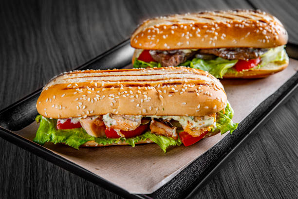 dwie długie kanapki podwodne z mięsem, serem, boczkiem, pomidorami, sałatą, ogórkami i cebulą na czarnym drewnianym tle stołu - panini sandwich zdjęcia i obrazy z banku zdjęć