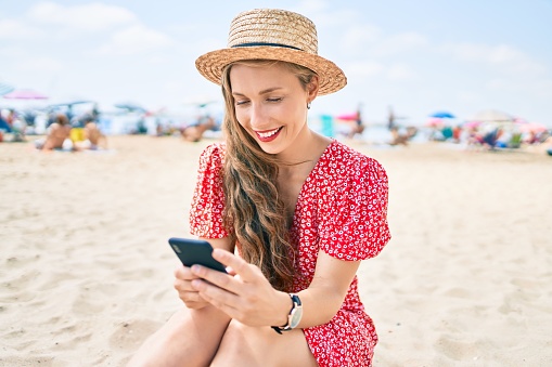 Young blonde woman on vacation using smartphone sitting on the sand at the beach
