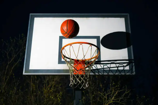 Photo of Street basketball ball falling into the hoop at night. Urban youth game. Close up of orange ball above the hoop net. Concept of success, scoring points and winning