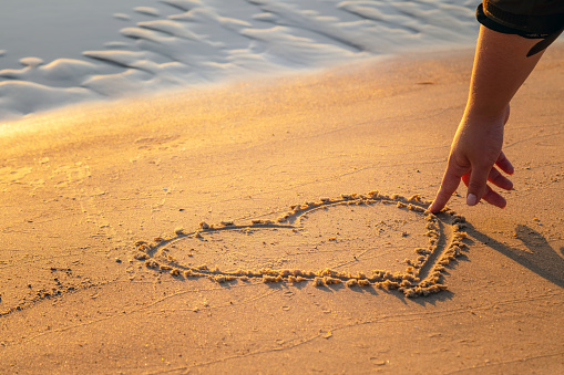 Close up view of woman hand drawing heart with finger on the sand at the beach. Romantic love symbol at tropical seashore at sunset