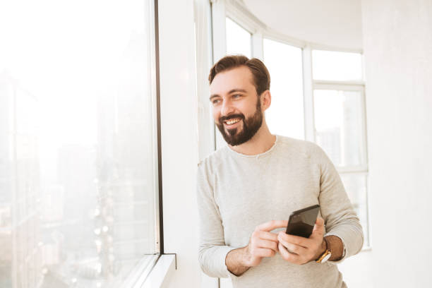 istock Communicative guy with in white shirt, looking through big window, while chatting or typing text message on mobile phone 1269328529