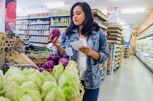 Shot of Southeast Asian woman picking up a red cabbage from retail display, while another hand holding a shopping list.