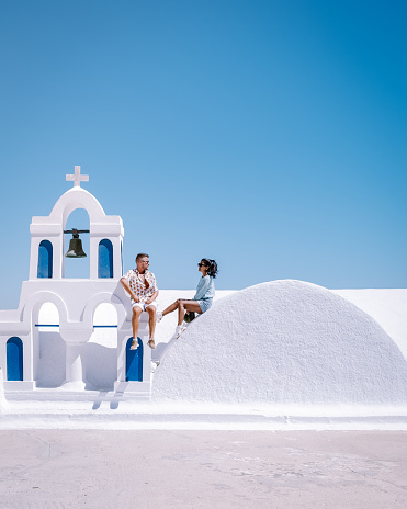 Santorini Greece, young couple on luxury vacation at the Island of Santorini watching sunrise by the blue dome church and whitewashed village of Oia Santorini Greece during sunrise during summer vacation, men and woman on holiday in Greece