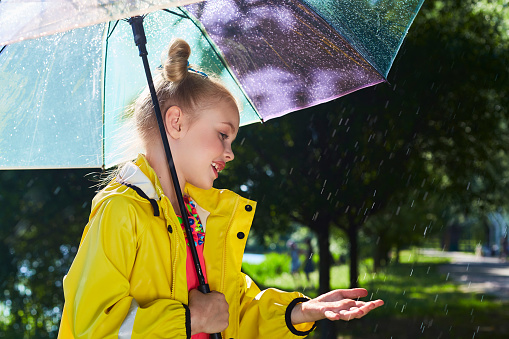horizontal frame. summer park is pouring rain and the sun is shining. beautiful blonde girl pulls her hand under the raindrops and smiles a toothless smile.