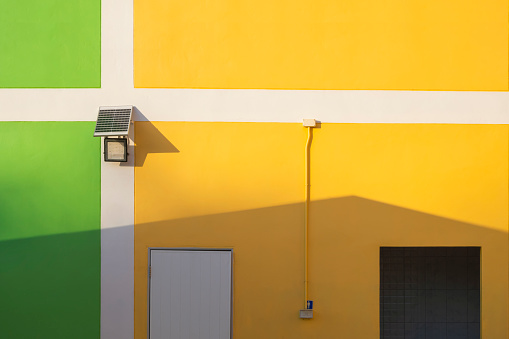 Sunlight and shadow on surface of small solar panel, spotlight, white door, restroom door with light switch box and electric pipeline on colorful building wall