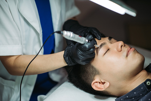 close up shot of male lying down on bed and receiving beautician using professional permanent makeup tattoo machine on eyebrow