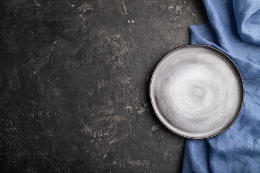 Empty gray ceramic plate on black concrete background and blue linen textile. Top view, copy space, flat lay.