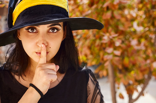 Portrait of beautiful female in witch hat sticking out her tongue and showing quiet sign. Attractive woman with little secrets showing silence gesture