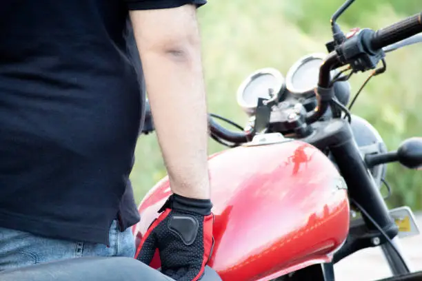 Photo of Young indian man wearing red riding gloves with protection while holding the handle bars of a motorcycle with a red tank