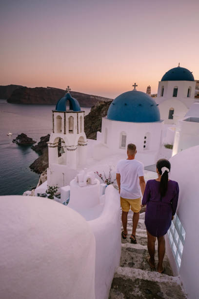 Santorini Greece, young couple on luxury vacation at the Island of Santorini watching sunrise by the blue dome church and whitewashed village of Oia Santorini Greece during sunrise Santorini Greece, young couple on luxury vacation at the Island of Santorini watching sunrise by the blue dome church and whitewashed village of Oia Santorini Greece during sunrise during summer vacation happy couple on vacation in santorini greece stock pictures, royalty-free photos & images