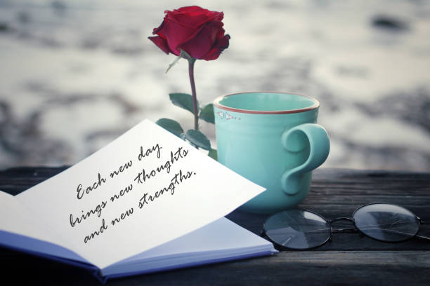 Inspirational quote written on a book paper - Each new day brings new thoughts, new strengths. With green cup of morning coffee or tea, notebook, glasses and red rose on the table on white background.