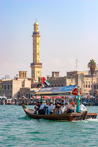 Dubai, United Arab Emirates - December 09, 2007: An Abra or Water Taxi is seen just departing from the piers of Bur Dubai for Deira, on the opposite side of the creek. On board are several passengers including a couple of tourists. In the background are the piers of the Bur Dubai Arba Station and some old buildings with Wind Towers. A single minaret of a mosque is also seen. Photo shot, from a boat on the creek, in the afternoon sunlight; horizontal format. Copy space.