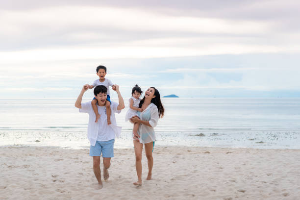 buone vacanze in famiglia asiatiche durante gioiosi padre, madre, figlio e figlia che camminano insieme lungo il mare estivo al tramonto. buon viaggio in famiglia in spiaggia in vacanza, estate e vacanza. - beach women joy sand foto e immagini stock