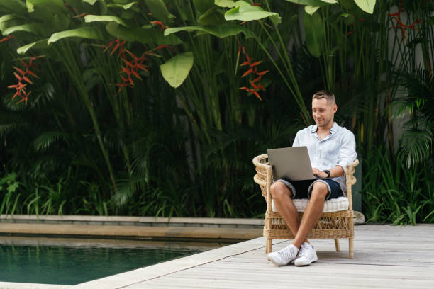 istock Young man using a laptop computer in a garden with a swimming pool. 1269444212