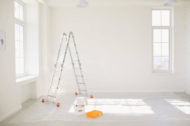 Painting in a white room with windows with a stepladder and paints on the floor.