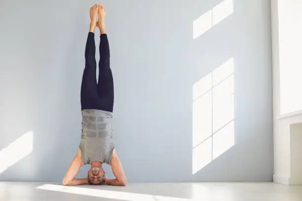 Healthy active mature male in activewear performing supported headstand near wall during morning yoga training in modern light studio