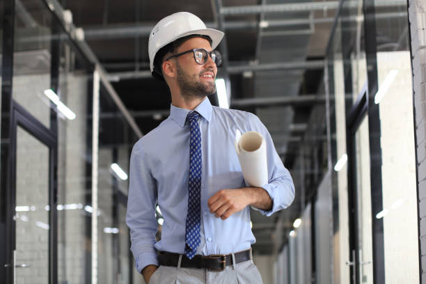 istock Confident young business man in hardhat holding blueprint and looking away while standing indoors. 1269484003