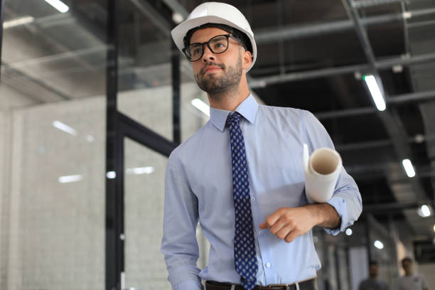 istock Confident young business man in hardhat holding blueprint and looking away while standing indoors. 1269486064