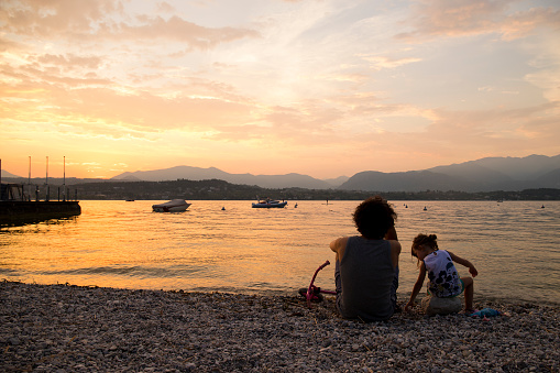 Young Father and little child daughter sitting on the lake beach at sunset in Manerba del Garda, Lombardy Italy. Back view.